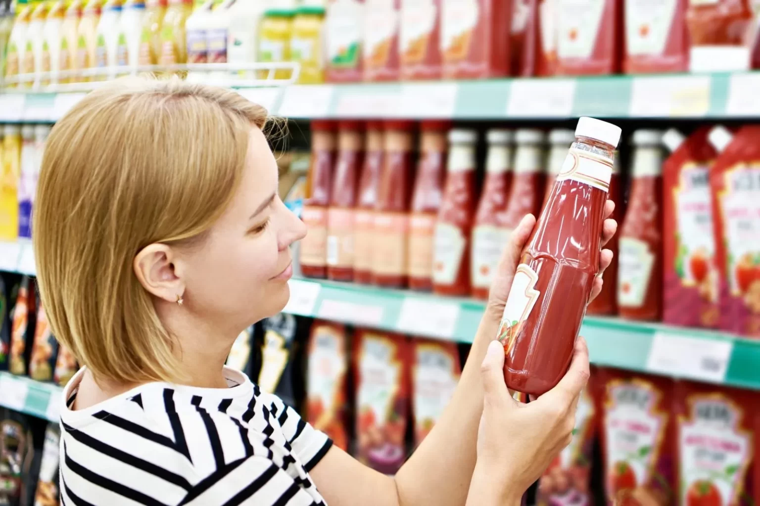 Woman choosing ketchup in supermarket aisle.